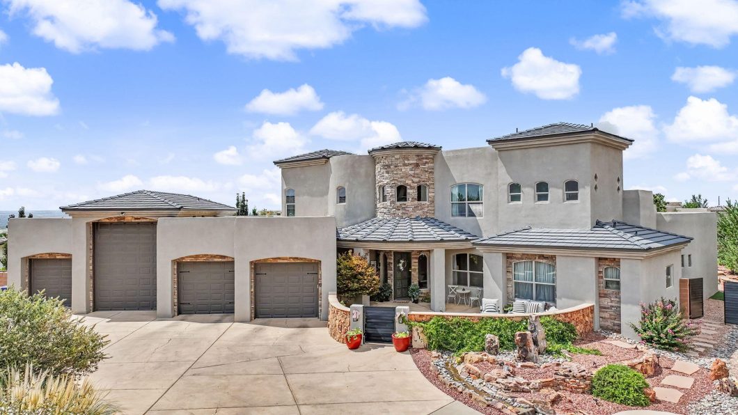 Modern two-story house with three attached garages, stone accents, and a front porch under a clear blue sky. Desert landscaping in the yard.